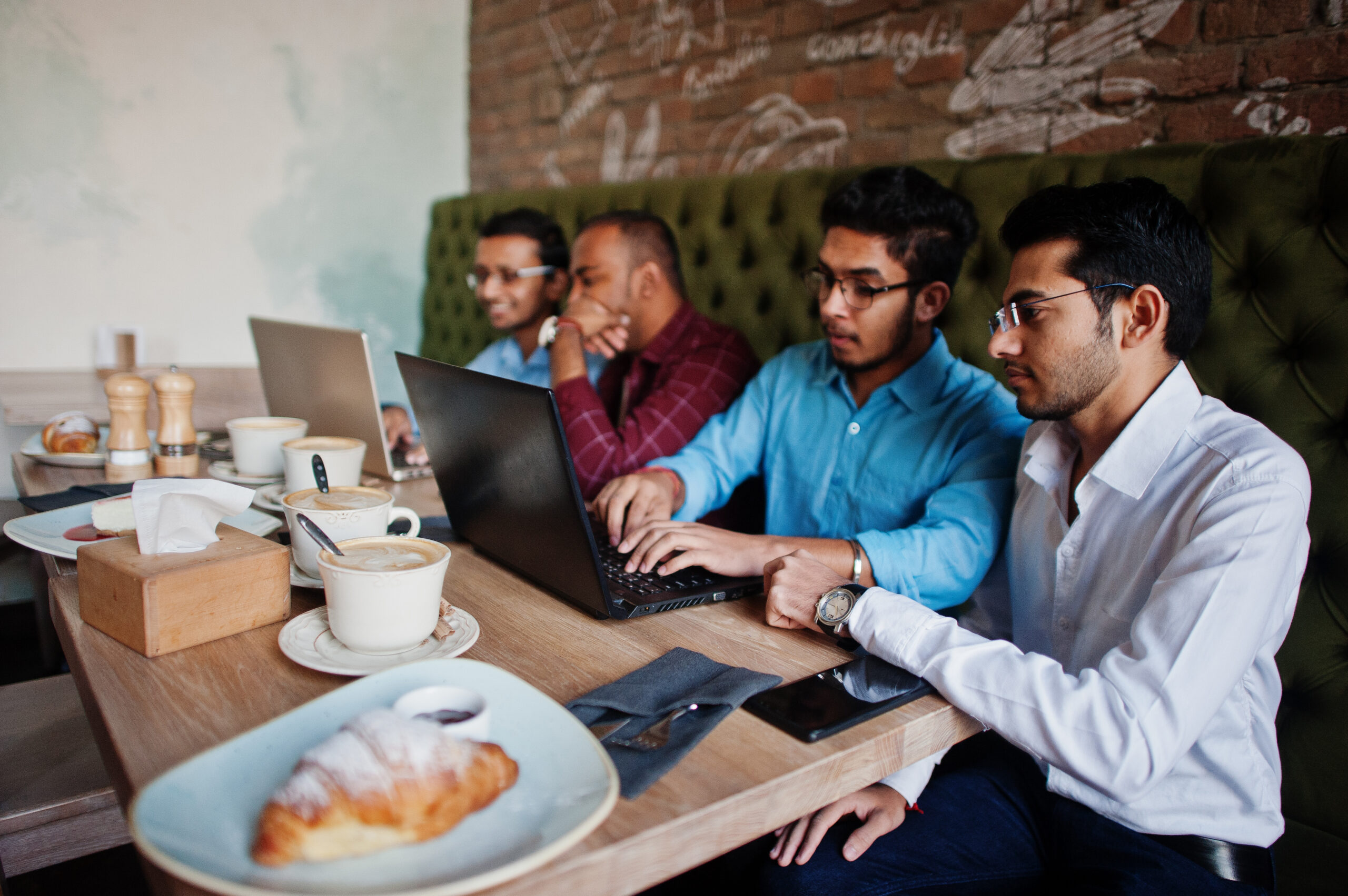 Group of four south asian men's posed at business meeting in cafe. Indians work with laptops together using various gadgets, having conversation.